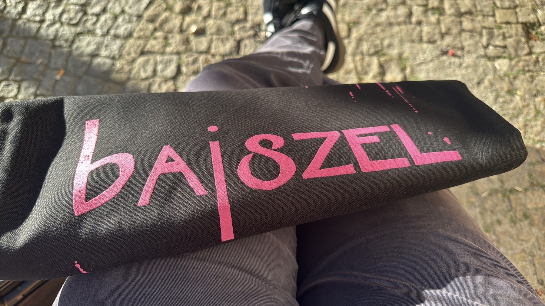 Ein schwarzel Beutel liegt auf meinen Beinen. Auf dem Beutel steht in Pink „bajszel“