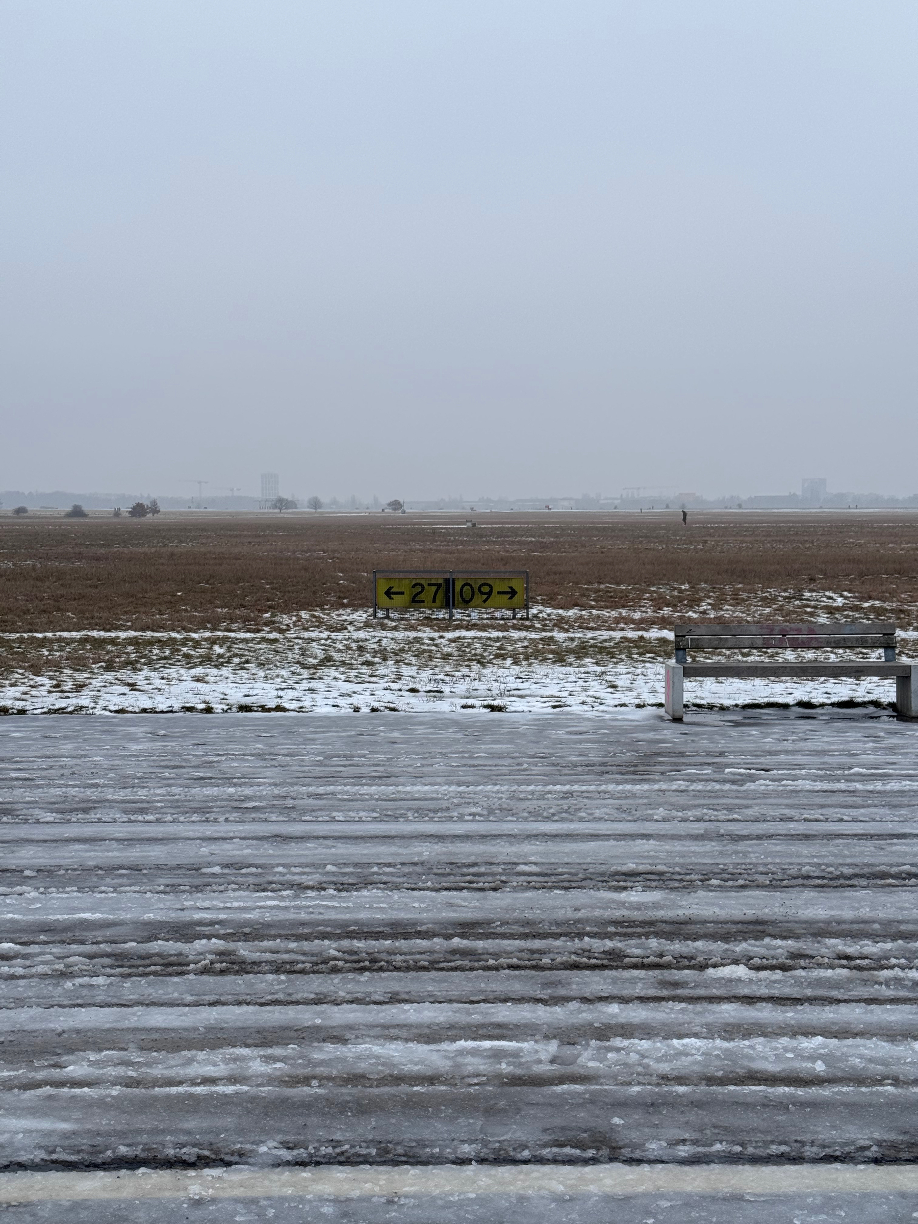 Ein tristes Foto vom Tempelhofer Feld: Eine Bank zwischen einem Flughafenschild und dem grauen Schneematsch vom Rollfeld.